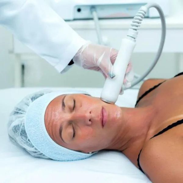 imagem de uma mulher deitada na maca de olhos fechados, enquanto a esteticista passa um equipamento de radiofrequência da HS Med no rosto da moça.
